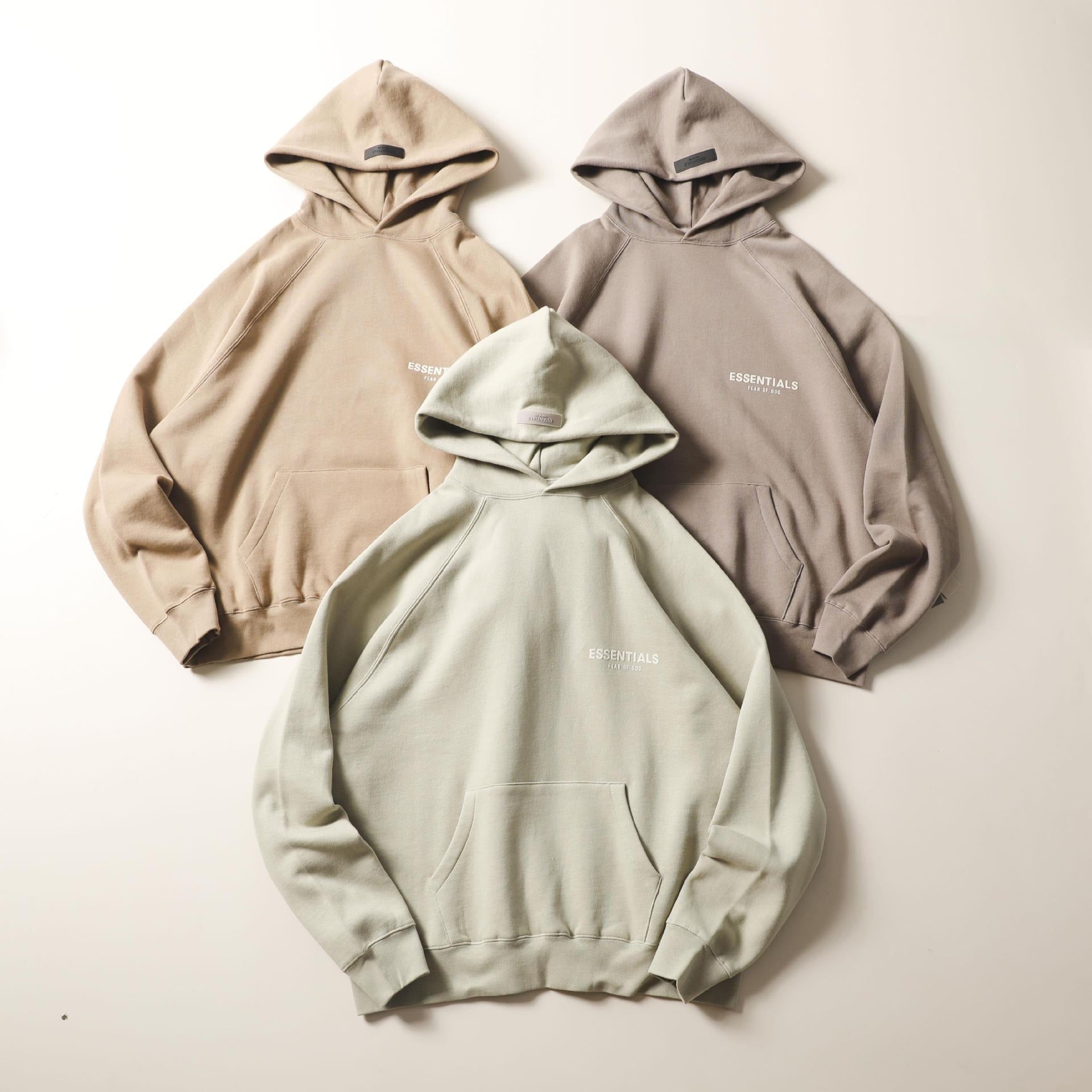 Beige and mint green hooded sweatshirts with "ESSENTIALS" text print on the chest, displayed against a plain white background.