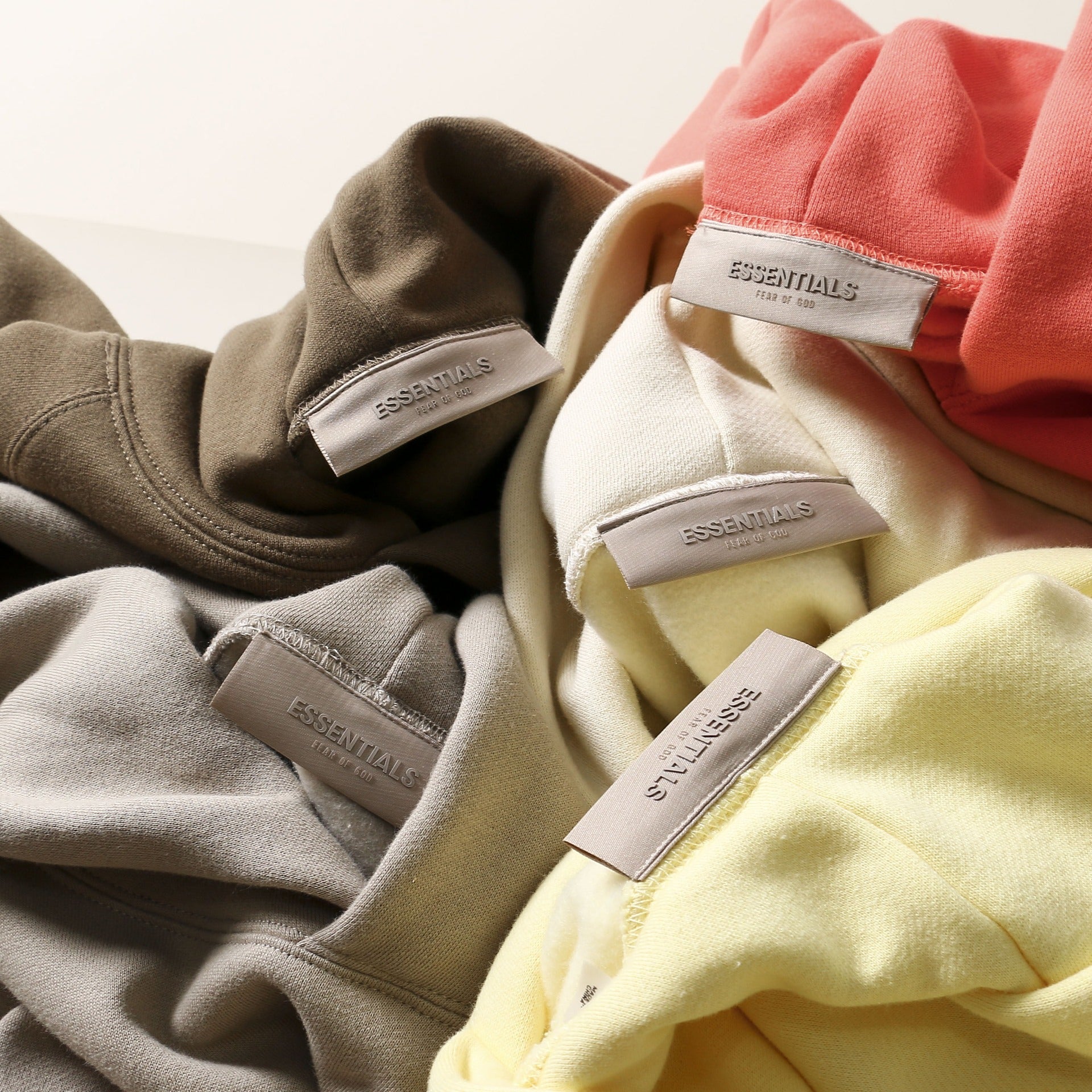 Cozy assortment of loose-fitting hoodies in various colors, including grey, white, and coral. The hoodies feature the "Essentials" brand label, indicating a casual, comfortable clothing line.